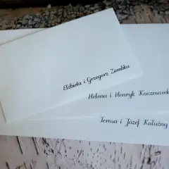 Białe koperty ślubne z elegancko wypisanymi nazwiskami gości na tle kory drzewa.