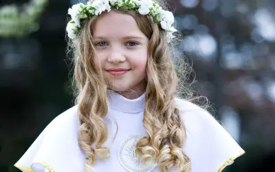Dziewczynka w wianku z białych kwiatów, ubrana w białą komżę, gotowa do przyjęcia Komunii Świętej. W tle słychać piękne pieśni komunijne.