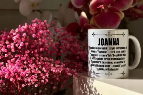 Kiedy imieniny Joanny? Na kubku z imieniem Joanna wyjaśnienie jego pochodzenia. W tle różowe kwiaty.