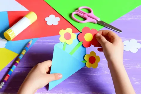 Dziecko tworzy laurkę dla nauczyciela na koniec roku. Kolorowe kwiatki z papieru wkładane są do niebieskiej koperty.
