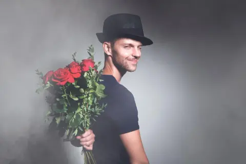 Elegancki mężczyzna w kapeluszu trzyma bukiet czerwonych róż za plecami. Idealny prezent na imieniny Marcina.