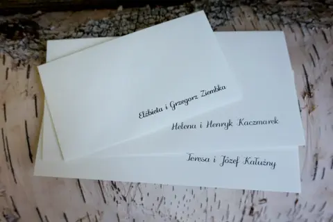 Białe koperty ślubne z elegancko wypisanymi nazwiskami gości na tle kory drzewa.