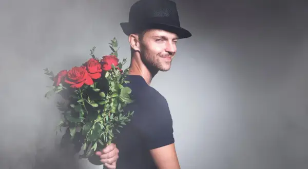 Elegancki mężczyzna w kapeluszu trzyma bukiet czerwonych róż za plecami. Idealny prezent na imieniny Marcina.