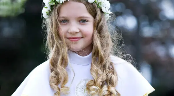 Dziewczynka w wianku z białych kwiatów, ubrana w białą komżę, gotowa do przyjęcia Komunii Świętej. W tle słychać piękne pieśni komunijne.