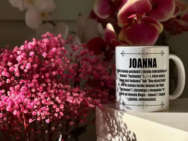 Kiedy imieniny Joanny? Na kubku z imieniem Joanna wyjaśnienie jego pochodzenia. W tle różowe kwiaty.