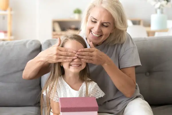 Babcia zasłania oczy wnuczce, która trzyma prezent. Radosna niespodzianka z okazji imienin Nikodema.